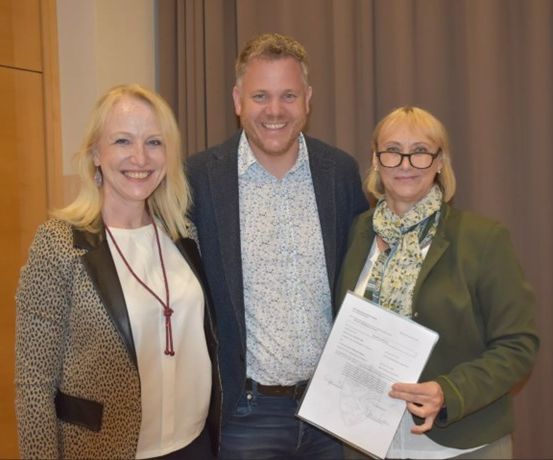 Drei Personen, zwei Frauen und ein Mann, stehen eng beieinander und lächeln für ein Foto. Die Frau auf der rechten Seite hält ein Zertifikat, und sie scheinen sich in einem Konferenzraum zu befinden.