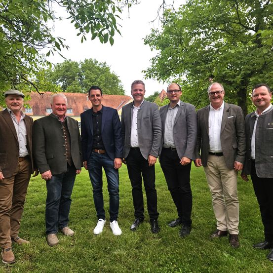Sieben Männer in Anzügen und Freizeitkleidung stehen in einem Park mit Bäumen und einem Haus im Hintergrund.
