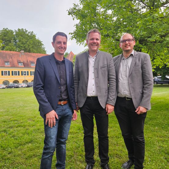 Drei Männer in Geschäftskleidung stehen in einem Park und lächeln vor einer Kamera vor einem Gebäude mit rotem Dach.