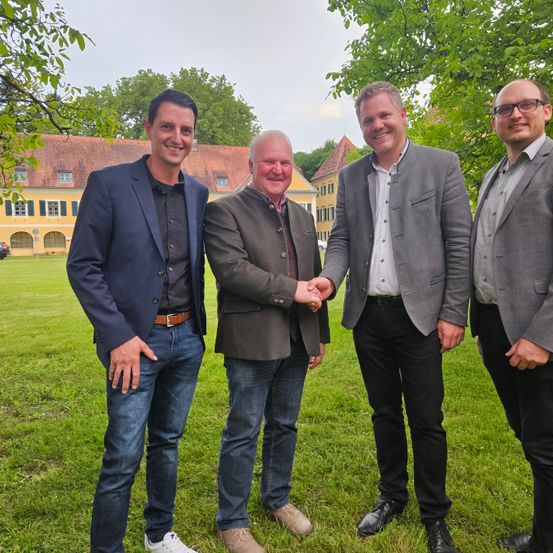 Vier Männer stehen in einer Reihe in einem Rasenbereich mit Bäumen, lächeln und schütteln Hände. Dahinter steht ein Gebäude mit einem braunen Dach und gelben Wänden.