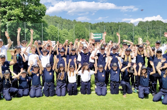Eine Gruppe von Personen in blauen Uniformen mit weißen Oberteilen posiert für ein Foto auf einem Rasenfeld. Sie heben ihre Hände, einige tragen Mützen und andere tragen Armbänder. Im Hintergrund befinden sich Bäume und ein Zaun.
