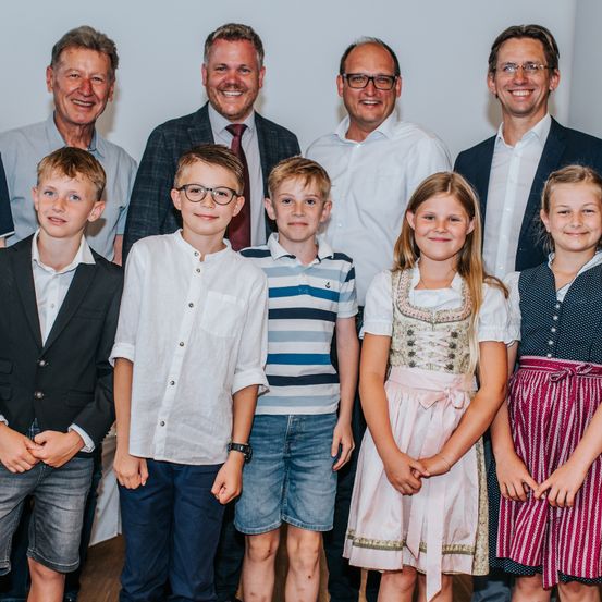 Eine Gruppe von Erwachsenen und Kindern posiert für ein Foto in einem Raum, sie tragen formelle und lässige Kleidung. Die Erwachsenen lächeln und die Kinder stehen eng beieinander.