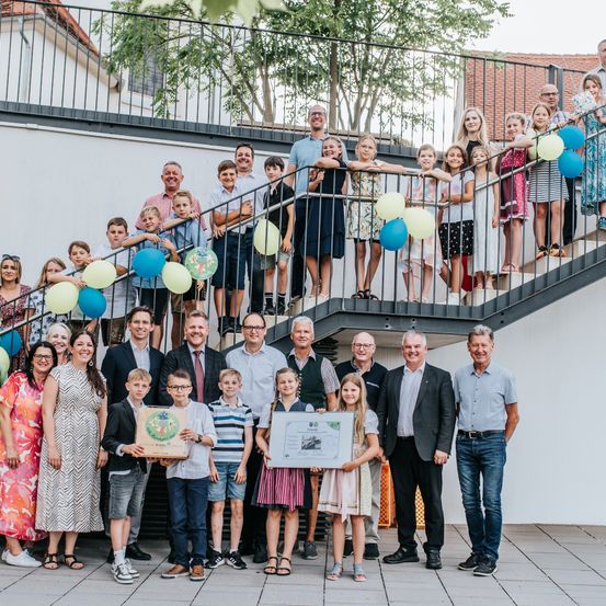 Eine Gruppe von Menschen posiert für ein Foto auf einer Treppe, hält Luftballons und Urkunden. Einige stehen auf der Treppe, andere unten, mit Gebäuden und Bäumen im Hintergrund.