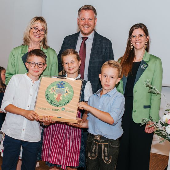 Eine Familie mit drei Kindern posiert für ein Foto, während sie eine Schachtel hält. Die Schachtel hat ein farbenfrohes Design mit einem Baum. Die Kinder tragen Freizeitkleidung. Die Erwachsenen sind in formeller Kleidung. Sie stehen in einem Raum mit weißen Wänden.