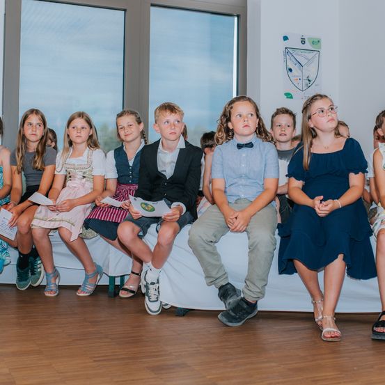 Mehrere Kinder sitzen auf einer Bank, jedes hält ein Stück Papier in der Hand, wahrscheinlich in einem Klassenzimmer mit großen Fenstern.