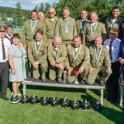 Eine Gruppe von Personen, gekleidet in grüne Uniformen und Medaillen, posiert für ein Foto auf einem Rasenfeld. Helme sind vor ihnen angeordnet. Dahinter steht ein Tisch mit Auszeichnungen.