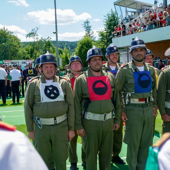 Soldaten in grünen Uniformen und Helmen stehen auf einem Feld und tragen Aufnäher mit Symbolen. Zuschauer beobachten von einer Tribüne oben.