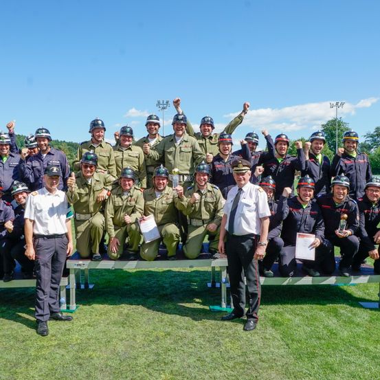 Eine Gruppe Feuerwehrleute in Uniformen und Helmen posiert auf einem Feld für ein Foto. Zwei Männer vorn halten Trophäen und Urkunden.