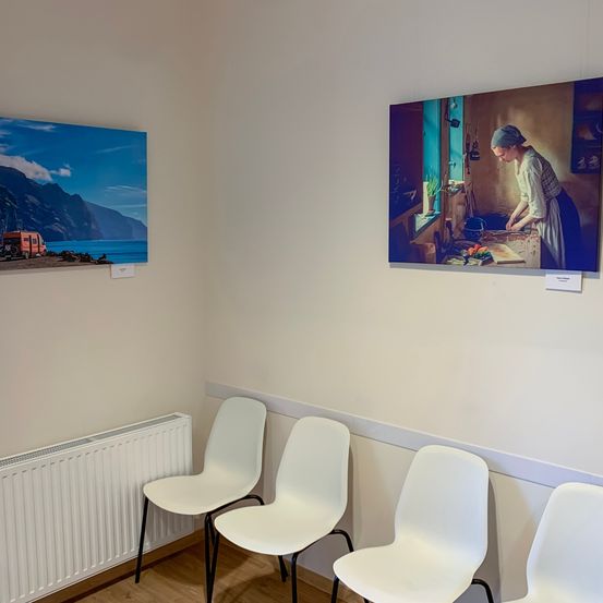 Ein Raum mit weißen Wänden und einem Holzboden ist mit einem Heizkörper und vier weißen Stühlen ausgestattet. An der Wand hängen zwei Gemälde, eines zeigt eine Frau beim Kochen und das andere eine Landschaft mit einem Van und Bergen.