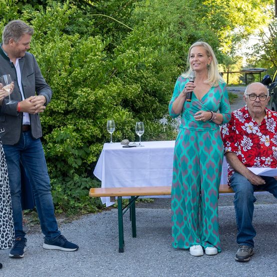 Eine Frau mit einem Mikrofon spricht bei einer Veranstaltung im Freien. Zwei Personen sitzen vor ihr auf einer Bank. Einer von ihnen hält ein Weinglas. Ein Tisch mit Weingläsern und einem Stein steht hinter ihnen.