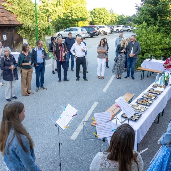 Eine Gruppe von Menschen, die möglicherweise an einer Veranstaltung teilnehmen, steht auf einem Parkplatz. Musikinstrumente und Notenblätter sind auf einem Tisch platziert. In der Nähe sind ein Gebäude mit einem grünen Pfosten und geparkte Autos zu sehen.