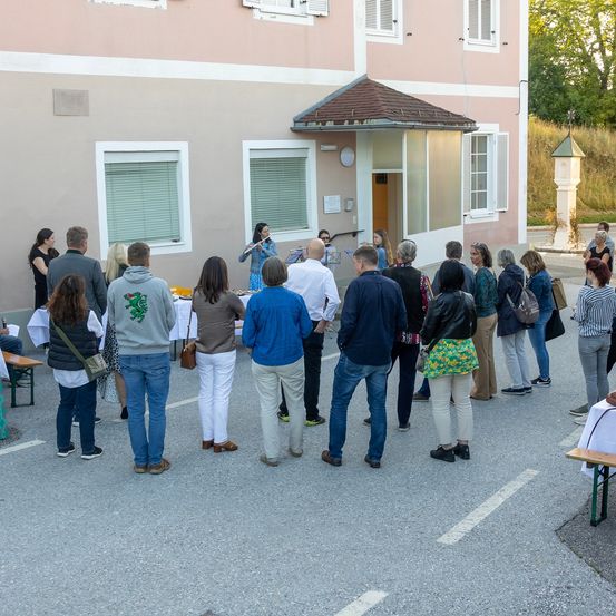 Eine Gruppe von Menschen steht vor einem rosa Gebäude mit Fenstern und einem kleinen Turm. Eine Frau spielt Flöte in der Nähe des Eingangs. Andere schauen zu, und einige tragen Turnschuhe.