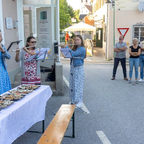 Vier Personen spielen Flöten in einer Außenumgebung. Sie stehen vor einem Gebäude mit einem Schild. Es gibt einen Tisch mit Essen darauf und eine Bank in der Nähe. Im Hintergrund befinden sich ein Verkehrsschild und einige stehende Personen.