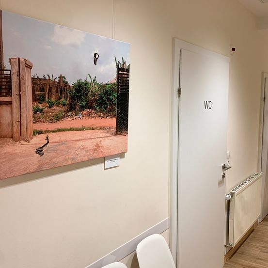 Ein großes Foto hängt an einer Wand neben einer weißen Tür mit der Aufschrift WC. Das Foto zeigt ein offenes Tor, das zu einem zerstörten Gebäude und einer Erdstraße führt.