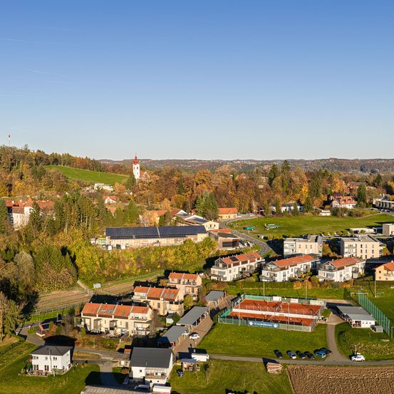 Eine Luftaufnahme eines Wohngebiets mit verschiedenen Häusern, einige mit Solarmodulen, umgeben von Bäumen und Grünflächen. Ein Turm ist in der Ferne sichtbar.