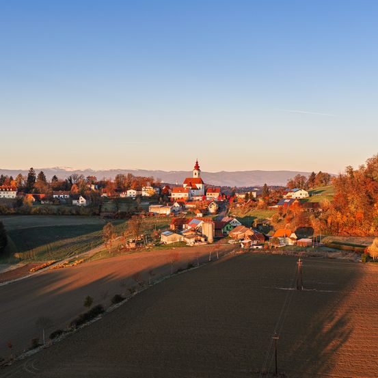 Eine Luftaufnahme eines Dorfes mit einer großen Kirche, umgeben von Feldern und Bergen unter einem blauen Himmel.