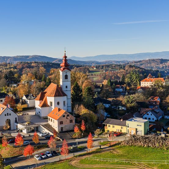 Ein Dorf mit Häusern, einer Kirche und Bergen in der Ferne. Autos sind vor der Kirche geparkt. Bäume mit roten Blättern sind verstreut.