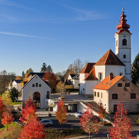 Auf einem Hügel steht eine Kirche mit rotem Dach und weißem Turm in einem Dorf. Herbstlaub in Rot und Gelb schmückt die Landschaft. Autos sind vor der Kirche geparkt.