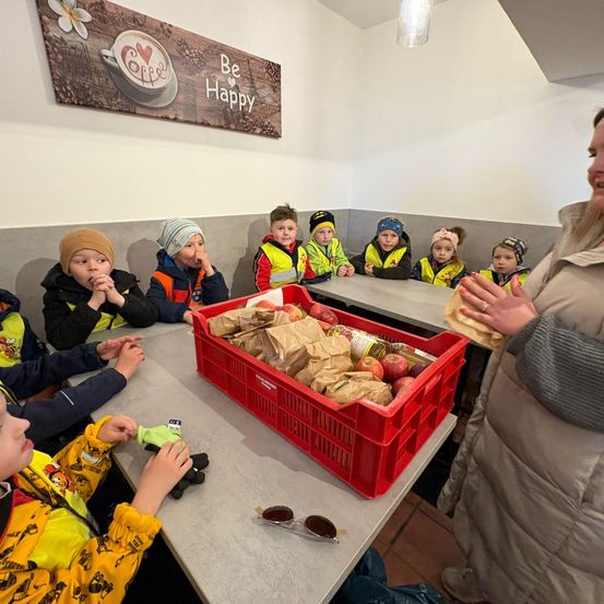 Eine Frau verteilt Essen an Kinder in gelben Westen am Tisch. Ein roter Kasten voller Äpfel steht auf dem Tisch. An der Wand befindet sich ein Schild mit der Aufschrift 'Sei glücklich'.
