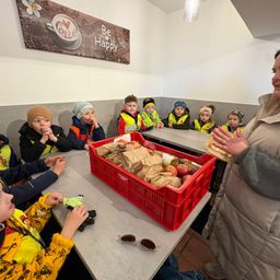 Eine Frau verteilt Essen an Kinder in gelben Westen am Tisch. Ein roter Kasten voller Äpfel steht auf dem Tisch. An der Wand befindet sich ein Schild mit der Aufschrift 'Sei glücklich'.