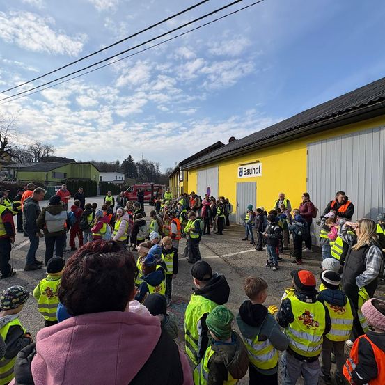 Viele Kinder in gelben Warnwesten versammeln sich vor einem Gebäude mit der Aufschrift 'Bauhof'. Einige Erwachsene sind anwesend, und ein Feuerwehrwagen ist in der Ferne zu sehen.