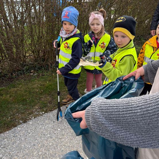 Eine Gruppe von Kindern in hellen Westen sammelt Müll in einem Park, wobei ein Kind einen Müllsack hält.