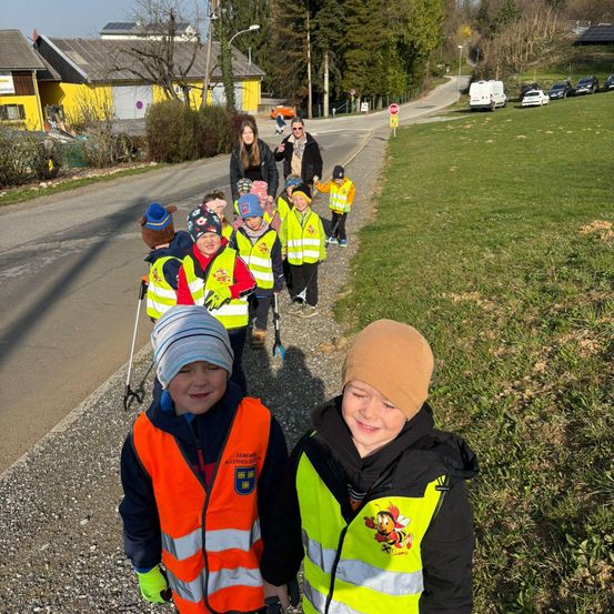 Kinder in gelben Warnwesten gehen auf der Straße, begleitet von Erwachsenen. Ein Grasbereich mit Bäumen ist an der Seite. Gebäude und Autos sind im Hintergrund sichtbar.
