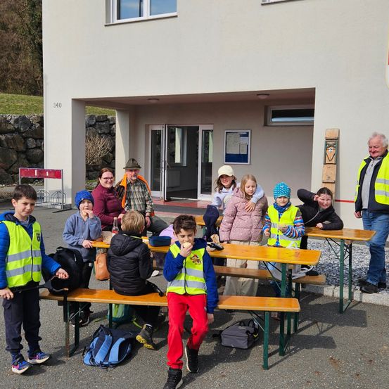 Eine Gruppe von Kindern und Erwachsenen, die reflektierende Westen tragen, sitzen an Tischen vor einem Gebäude. Einige essen, während andere lächeln. Ein Mann steht in der Nähe des Gebäudes.