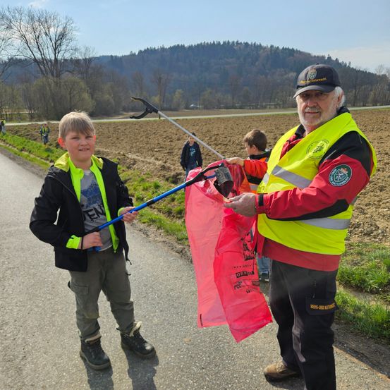 Ein älterer Mann und ein junger Junge in Jacken sammeln Müll am Straßenrand. Der Junge hält einen Stock mit einem Netz, und der Mann hält einen Plastiksack. Dahinter ein Feld mit anderen Menschen, die Müll sammeln.