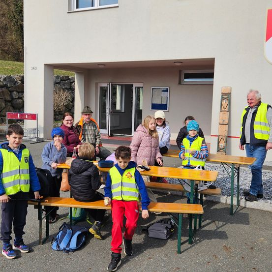 Eine Gruppe von Kindern und Erwachsenen, die Sicherheitswesten tragen, versammelt sich um einen langen Tisch vor einem Gebäude. Einige sitzen auf Bänken, andere stehen, mit Taschen und einem Mann in einer reflektierenden Weste in der Nähe.