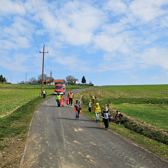 Eine Gruppe von Kindern und Erwachsenen in gelben Westen läuft auf einer Straße. Die Straße ist von grünen Feldern umgeben. Ein rotes Fahrzeug ist am Straßenrand geparkt.