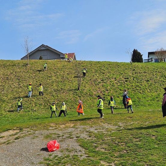 Eine Gruppe von Kindern in Warnwesten geht auf einem grasigen Hügel. Einige haben Handschuhe. In der Ferne sind Häuser zu sehen.
