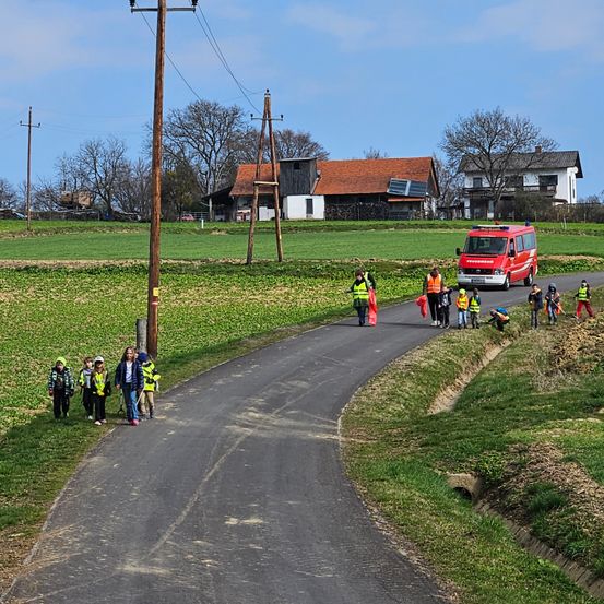 Kinder in Warnwesten laufen an einer Straße mit einem roten Notfallfahrzeug. Die Straße wird von Feldern und Häusern flankiert, wobei einige Häuser Solarmodule auf dem Dach haben.