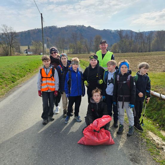Eine Gruppe von Kindern und Erwachsenen posiert für ein Foto am Straßenrand, mit Müllsäcken in den Händen. Sie tragen Warnwesten und Handschuhe, wobei eine Frau und ein Mann an den Seiten stehen. Berge und ein grünes Feld sind im Hintergrund.