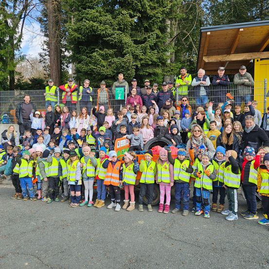 Eine Gruppe von Kindern und Erwachsenen in Warnwesten posiert für ein Foto vor einem Gebäude mit Zaun, Bäumen und einem Fahrzeug.
