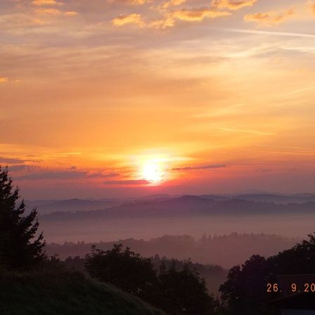 Sonnenuntergang über einer nebligen Landschaft mit Bergen im Hintergrund. Der Himmel ist orange und pink, mit einem Datum vom 26. September sichtbar.