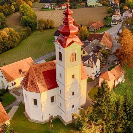 Luftaufnahme einer Kirche mit rotem Turm und gelben Wänden, eingebettet in eine ländliche Landschaft mit grünen Feldern und braunen Dächern.
