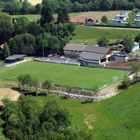 Luftaufnahme eines Fußballfeldes mit einem großen Gebäude und einem Parkplatz in einem ländlichen Gebiet, umgeben von Bäumen und Häusern.