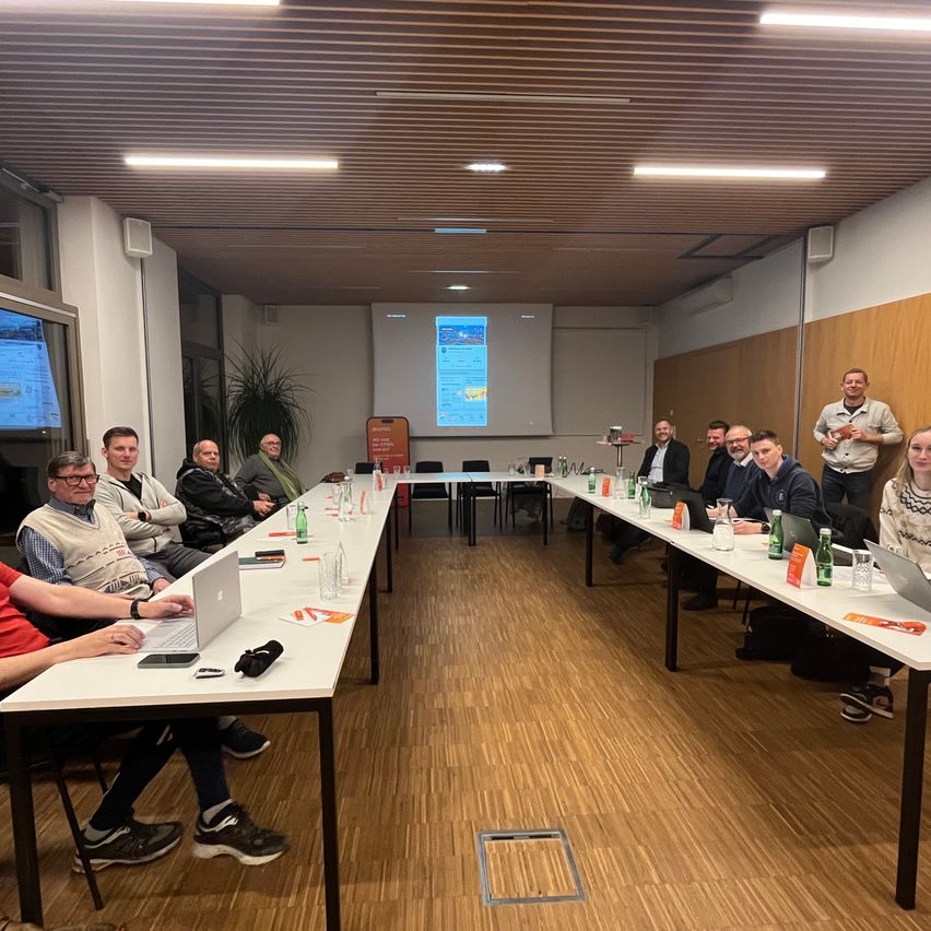 Eine Gruppe von Menschen sitzt in einem Besprechungsraum um Tische herum. Auf den Tischen sind Laptops, Wasserflaschen und Papiere. Eine Projektionsleinwand ist an der Wand, und ein Mann steht davor.