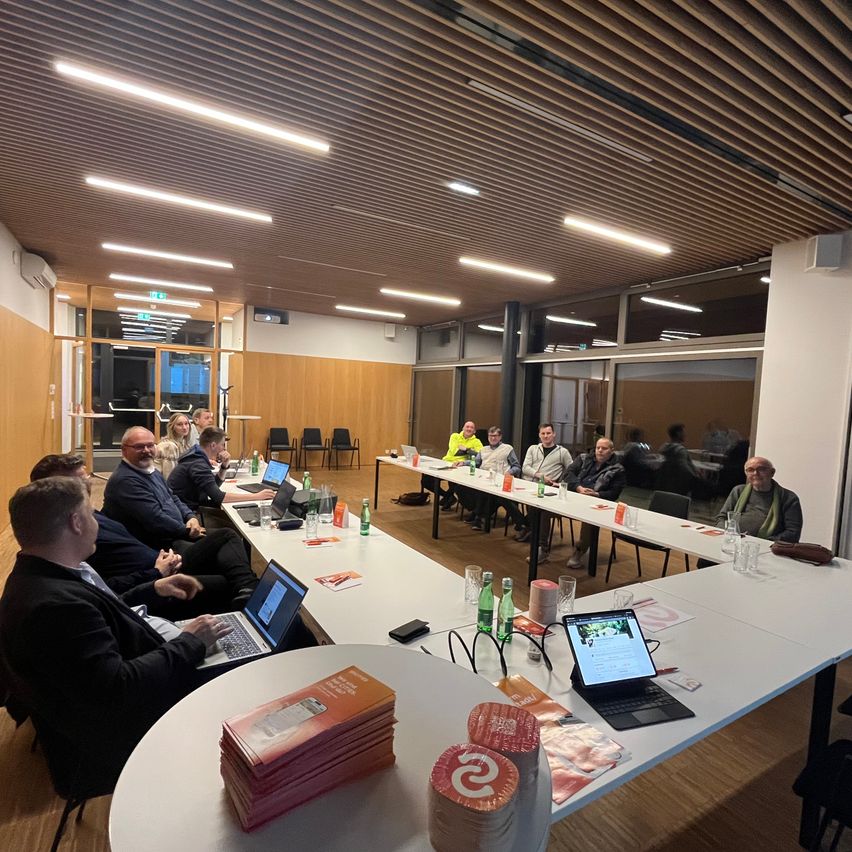 Eine Gruppe von Menschen sitzt um einen langen Tisch in einem Besprechungsraum herum und führt eine Diskussion. Laptops, Bücher und Tassen stehen auf dem Tisch. Der Raum hat große Fenster, Holzwände und eine Decke mit Beleuchtung.