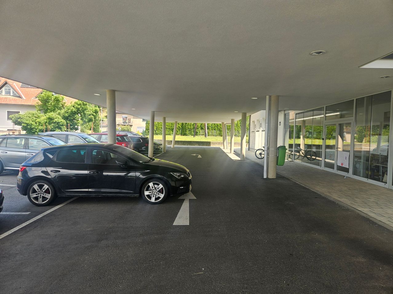 Ein schwarzes Auto ist auf einem Parkplatz mit weißen Markierungen geparkt, unter einem überdachten Bereich mit Säulen und Glaswänden.