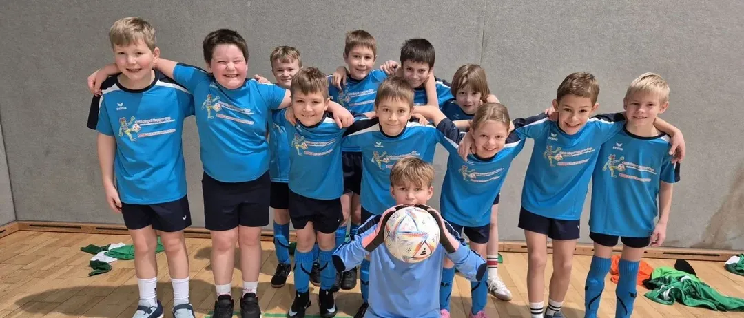 Eine Gruppe junger Jungen in blauen Trikots steht und kniet auf einem Holzboden, hält einen Fußball und posiert für ein Foto.