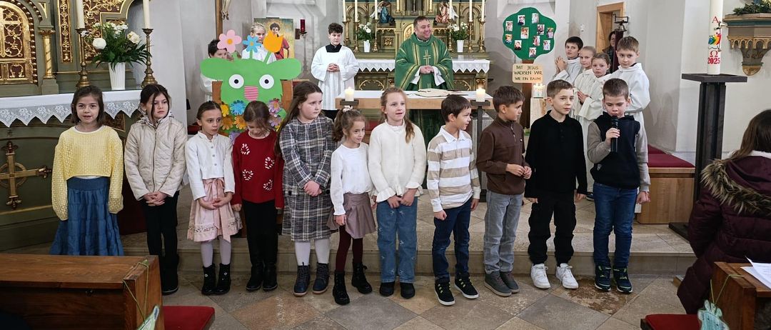 Eine Gruppe von Kindern steht in einer Kirche und schaut auf einen Priester, der am Altar steht. Der Altar ist mit Kerzen und Blumen geschmückt, und der Priester lächelt. Die Kinder tragen Winterkleidung, und einige halten ein Mikrofon.