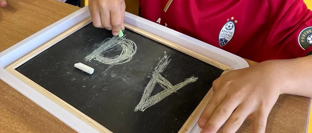 Ein kleiner Junge in einem roten Trikot schreibt mit Kreide auf einem Schultafel im Klassenzimmer. Ein weiteres Kind steht hinter ihm, und im Hintergrund befinden sich Lagerboxen.