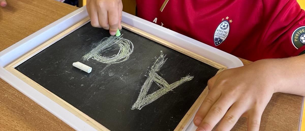 Ein kleiner Junge in einem roten Trikot schreibt mit Kreide auf einem Schultafel im Klassenzimmer. Ein weiteres Kind steht hinter ihm, und im Hintergrund befinden sich Lagerboxen.