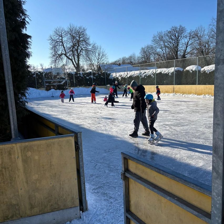 Mehrere Menschen, darunter Kinder, skaten auf einer Eisbahn im Freien, umgeben von Schnee und Bäumen. Sie tragen Winterkleidung und Helme.