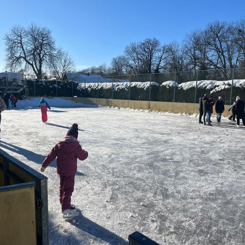 Mehrere Menschen skaten auf einer Eisbahn im Freien. Ein Kind in einem rosa Outfit fährt in Richtung Kamera. Im Hintergrund sind schneebedeckte Büsche und ein Zaun.