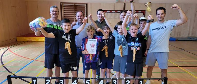Gruppenfoto von jungen Athleten und Trainern in einer Turnhalle. Sie halten Pokale und Medaillen. Nummerierte Kürbisse stehen vor ihnen auf einem Gestell.