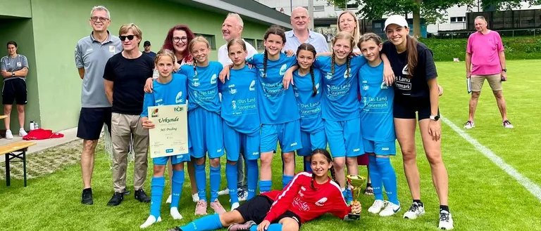 Eine Fußballmannschaft posiert auf einem Feld mit Erwachsenen für ein Foto. Ein Mädchen hält einen Pokal und andere halten Urkunden. Sie tragen blaue und rote Trikots. Ein Gebäude mit Balkon steht hinter ihnen.