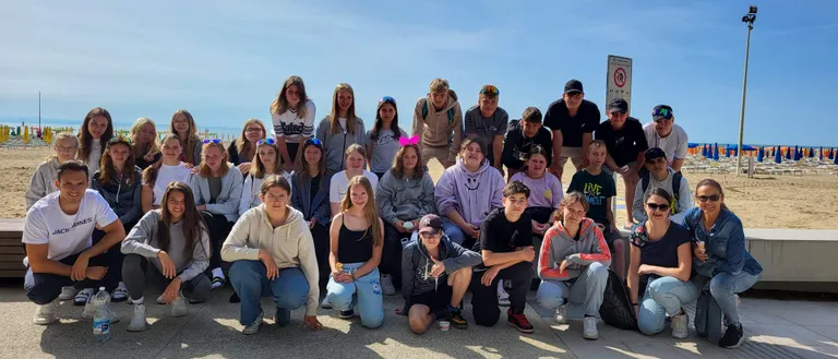 Eine große Gruppe von Menschen posiert für ein Foto am Strand, mit klarem blauem Himmel und einem Schild im Hintergrund.